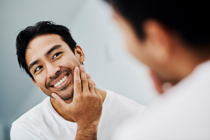 Handsome male touching his healthy face in the mirror after a skincare treatment. Confident man feeling fresh and attractive from a beauty routine. Portrait of a guy looking at his skin. LaseMD Ultra In New Jersey | Geria Dermatology